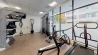 This fitness center is designed with both functionality and aesthetics in mind, offering a well-organized layout across a single level. The space is defined by a wall of floor-to-ceiling windows along one side, flooding the interior with natural light and providing views of the surrounding streetscape and greenery. Light wood-tone flooring runs throughout, complementing the clean white walls and drop-panel ceiling with recessed and flush-mounted lighting fixtures.

The cardio zone features a row of Matrix treadmills and stationary bikes positioned along the windowed wall, making workouts visually engaging. A large wall-mounted mirror extends across one end of the room, enhancing the sense of depth and allowing users to monitor their form. The strength training area is equipped with a professional-grade functional cable trainer, a TRX suspension training system mounted to the wall, and additional resistance equipment, providing a comprehensive range of workout options.

A wall-mounted television is installed in the training area for entertainment or instruction. The space also includes glass-paneled interior doors, contributing to an open, airy feel while maintaining distinct zones within the fitness center. Overall, the design strikes a balance between a clean, modern aesthetic and practical, high-quality fitness amenities.