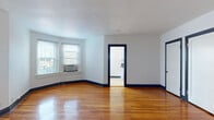 This single-floor residence offers a well-organized layout connected by a central hallway with polished hardwood flooring and ceiling-mounted lighting. A consistent design language runs throughout the space, characterized by crisp white walls, bold navy door and baseboard trim, and warm honey-toned hardwood floors that add richness to every room.

The spacious living area is bright and open, featuring large windows with window-unit air conditioning and generous wall space. An adjacent room offers flexibility as a dining area or second living space, also bathed in natural light through dual windows and equipped with a ceiling fan.

The galley-style kitchen is efficiently designed with white cabinetry lining both walls, dark countertops, subway tile backsplash, and black stainless appliances including a gas range and refrigerator. Tile flooring completes the practical aesthetic.

The bedrooms are comfortably sized, each with hardwood floors and navy-trimmed doorways. One bedroom features a ceiling fan, dual windows, and a classic cast-iron radiator for supplemental heating. Another includes a mirrored closet door, adding visual depth.

The bathroom is clean and functional, featuring tile flooring, a pedestal-style vanity, a window for natural light, and a cast-iron radiator. The overall ambiance is tidy, classic, and move-in ready.