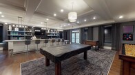 Located at 2470 Merchant Street in Frederick, Maryland, this single-floor amenity space is designed with a sophisticated, contemporary aesthetic that balances comfort and entertainment. The interior is finished with warm hardwood flooring throughout, complemented by rich gray wall paneling, crisp white crown molding, and tray ceilings adorned with recessed lighting and statement pendant fixtures.

The game room is a central highlight, housing a full-size billiards table and a foosball table set against a backdrop of wall-mounted televisions. Floor-to-ceiling drapes in deep charcoal frame the windows, lending a refined, moody ambiance. An adjacent lounge area offers generous sectional seating alongside cognac leather accent chairs, all anchored by layered area rugs beneath modern circular halo pendant lights — creating an inviting space for relaxed gathering.

The wet bar and kitchenette area features white cabinetry, open floating shelves set against a dark subway tile backsplash, and a large quartz-topped island with bar seating. Built-in appliances and a sink make this area fully functional for entertaining. Large windows with Venetian blinds and floor-length curtains provide natural light while maintaining privacy. French glass doors connect the space to adjacent areas, enhancing the open, airy flow throughout.