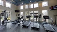 This fitness center is designed to provide a bright and motivating environment for exercise. The space features soaring ceilings accented by a deep navy upper wall and ceiling treatment, creating a bold architectural contrast against the crisp white lower walls. Large, floor-to-ceiling windows line the main wall, flooding the room with natural light and offering clear views of the surrounding landscaped community — giving the space an open, airy feel despite its focused function. The flooring consists of durable dark slate-toned tiles, practical and well-suited for a high-traffic fitness environment. The cardio equipment lineup includes multiple treadmills, an elliptical trainer, a stationary bike, and a stair climber, providing a comprehensive range of aerobic workout options. A wall-mounted flat-screen television is positioned for easy viewing during workouts. Recessed lighting and a ceiling fan complement the natural light, ensuring the space remains well-lit and comfortable throughout the day. The layout is thoughtfully arranged to maximize usable floor space while maintaining clear sightlines to the outdoors, contributing to an uplifting and energizing atmosphere for residents.