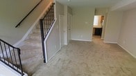 This two-story home offers a thoughtfully laid-out interior designed for comfortable everyday living. The main level greets you with a spacious open living area finished with wall-to-wall carpeting in a neutral beige tone, creating a warm and inviting atmosphere throughout. Smooth, light-toned walls complement the clean white baseboards, lending the space a bright and cohesive look.

A prominent staircase with dark metal balusters and a contrasting dark handrail serves as a key architectural feature, providing a stylish transition between the two floors while adding visual interest to the main living area. Underneath the staircase, the space is utilized efficiently, and a pair of interior doors nearby suggest convenient closet or storage access.

Adjacent to the living area, the kitchen is visible through an open passageway and features dark cabinetry paired with light countertops, creating a classic contrast. A window with vertical blinds allows natural light to filter into the kitchen area, enhancing the overall brightness of the home.

The two-floor layout offers a clear separation between the living and private spaces of the home, making it both practical and functional. The consistent use of neutral tones, carpeted flooring, and clean architectural lines gives this home a tidy, move-in-ready character throughout.