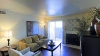 This single-story space features a thoughtfully arranged living area that serves as the centerpiece of the home. The living room is finished with warm hardwood flooring and neutral-toned walls that create a bright, inviting atmosphere. A textured ceiling adds subtle architectural character to the room.

A standout feature is the fireplace, positioned along one wall and framed with a clean, classic surround, providing both a functional heating element and a natural focal point for the space. Decorative ironwork wall art adorns the wall nearby, adding visual interest and a touch of elegance.

Natural light flows generously into the living area through a sliding glass door fitted with vertical blinds, offering access to an outdoor area and views of the exterior surroundings. This connection to the outdoors enhances the overall sense of openness within the space.

The living room comfortably accommodates a full seating arrangement, with ample room for a sofa, accent chair, and coffee table. A floor lamp supplements the natural light, ensuring the space remains well-lit throughout the day. Overall, the space presents a functional and comfortable single-floor layout with warm, welcoming finishes.