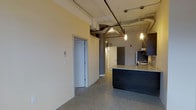 This single-floor space showcases a compelling blend of industrial and contemporary design elements. The open layout is defined by polished concrete floors that extend throughout, lending a clean, urban aesthetic underfoot. Exposed ductwork and piping run along the ceiling, complementing the industrial character of the space while contributing to its loft-like atmosphere. A cylindrical structural column adds an architectural detail that reinforces the building's raw, converted character.

The kitchen is a standout feature, anchored by a substantial island with a light granite countertop and dark cabinetry below. Pendant lights hang above the island, providing focused task lighting with a modern touch. Dark upper cabinetry lines the kitchen wall, paired with stainless steel appliances and a range hood, creating a cohesive, functional workspace.

The open corridor beyond leads to additional rooms, with neutral-toned walls and white-painted door frames offering a clean contrast to the industrial ceiling details. The overall ambiance is modern and utilitarian, with carefully chosen finishes that balance raw architectural elements with refined touches throughout.