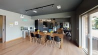 Located at 15 Huntington Cir in Naperville, IL, this single-floor space blends industrial design with modern comfort throughout its thoughtfully arranged layout. The main gathering area features light hardwood flooring, exposed ductwork, and a dark ceiling with black structural beams, creating a striking industrial aesthetic. A large wall-mounted TV and a mix of seating — including a rich leather sofa, accent chairs, and a patterned area rug — anchor the lounge zone, while a pendant globe chandelier adds a warm, contemporary focal point. Adjacent to the lounge, a generous communal dining table surrounded by classic Eames-style chairs provides ample space for group gatherings, alongside additional round dining tables for smaller groups. A dedicated workstation area with computers is also available. The fully appointed kitchen is a standout feature, showcasing dark cabinetry, a textured stone tile backsplash, warm wood accents, white quartz countertops, a stainless steel refrigerator, and pendant lighting — all combining for a refined yet functional culinary space. The fitness room is well-equipped with strength training machines, a stationary bike, full-wall mirrors, and natural light from street-facing windows. Large sliding glass doors in the lounge open directly to an outdoor patio, bringing in abundant natural light and seamless access to exterior seating surrounded by greenery.