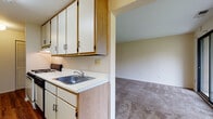 Located at 16 Brandywine in Amherst, Massachusetts, this single-floor residence offers a practical and functional layout suited for comfortable everyday living. The galley-style kitchen is efficiently designed with ample white cabinetry accented by warm wood trim, white laminate countertops, a gas range, a double-door refrigerator, and a stainless steel sink. Rich hardwood-style flooring runs throughout the kitchen, adding warmth and easy maintenance to the space. The living area is generously sized and bathed in natural light, thanks to a large sliding glass door that opens to a view of lush green outdoor surroundings, creating a seamless indoor-outdoor connection. Neutral-toned carpet extends through the living room and into the bedroom areas, providing a soft and comfortable underfoot feel. The bedroom features louvered closet doors offering ample storage, along with a window that allows natural light to filter in. A dedicated study or utility nook with a built-in desk adds functional versatility. The full bathroom includes a tub-shower combination set within white subway tile surround, a wood-toned vanity with an integrated sink, and classic diamond-patterned floor tile detailing. Overall, the space presents a clean, neutral palette throughout.