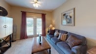 This single-floor residence offers a cohesive layout with warm, neutral tones carried consistently throughout each space. The living room is carpeted and features French doors that flood the area with natural light and provide outdoor views, complemented by a ceiling fan and decorative wall shelving. An adjacent dining area accommodates a full dining table beneath an elegant chandelier, with decorative accents and a tall indoor plant adding a welcoming atmosphere.

The galley-style kitchen is efficiently designed with warm wood cabinetry lining both sides, light-toned countertops, tile flooring, and black appliances including a range, microwave, refrigerator, and dishwasher. Ample storage is provided by the generous cabinet configuration.

The bedroom is carpeted and features a window with blinds and drape panels that allow for natural light control. A sitting area within the room adds a functional touch. The bathroom includes a single-sink vanity with wood cabinetry, a tub-shower combination with tile surround, and tile flooring, all finished with brushed nickel fixtures. Overall, the space presents a practical and cohesive design with consistent warm finishes.