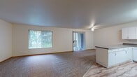 Located at 1616 NE 16th Way in Gresham, Oregon, this single-floor residence offers a clean, functional layout with comfortable living spaces throughout. The expansive living room is a standout feature, with plush carpeting, neutral white walls, and abundant natural light streaming in through a large picture window and a sliding glass door fitted with vertical blinds. A ceiling fan adds comfort, while the sliding door provides direct access to the outdoors and views of surrounding greenery. The kitchen is well-equipped with white cabinetry offering ample storage, dark laminate countertops, a double sink, a white electric range, a dishwasher, and light wood-look flooring that adds warmth to the space. A breakfast bar-style peninsula separates the kitchen from the adjoining areas. The bedrooms are carpeted and offer natural light through vertical-blind windows, with at least one bedroom featuring a sliding wood-door closet with built-in shelving. The bathrooms include light wood-look flooring, white vanity cabinetry with neutral countertops, wall mirrors, and grab bars. One bathroom includes a full tub and shower combination. Overall, the space presents a tidy, practical design suited for comfortable everyday living.