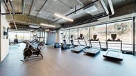 Located at 127 W Pomona St in Monrovia, CA, this single-floor fitness facility offers a generously sized, well-equipped exercise space divided into two distinct exercise rooms. The design embraces an industrial-modern aesthetic, characterized by exposed concrete ceilings with visible ductwork and piping, durable rubber flooring, and floor-to-ceiling windows that flood the space with natural light and provide open views of the surrounding landscape.

The primary exercise room is expansive and thoughtfully zoned for multiple training disciplines. A dedicated cardio section lines the window wall, featuring treadmills, ellipticals, stair climbers, and stationary bikes. The strength training area is equipped with cable machines, selectorized weight stations, a squat rack, and a full dumbbell rack with wall mirrors. A functional training zone includes rowing machines, a punching bag, TRX suspension trainers, and open floor space for dynamic movement.

The second exercise room offers a more intimate setting with accent walls in bold yellow, rubber-tiled flooring, and storage racks stocked with medicine balls, kettlebells, and stability balls. A separate studio area features a wall-mounted display screen, open mat space ideal for yoga or group classes, and acoustic slatted wall panels for a refined finish. A water station is conveniently accessible throughout the facility.