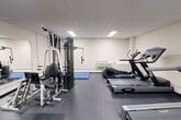 This fitness room at 935 Dundas St E in Mississauga, Ontario offers a practical and functional exercise space designed to accommodate a variety of workout routines. The room features a drop-panel ceiling with recessed fluorescent lighting that ensures consistent, even illumination throughout the space. Dark rubber flooring provides a durable, non-slip surface suited for active use.

The cardio section is well-appointed, featuring multiple treadmills, an elliptical trainer, and a stationary bike, giving users a range of aerobic options. A wall-mounted flat-screen television is positioned within view of the cardio equipment for entertainment during workouts. On the strength training side, a multi-station cable machine offers versatility for full-body resistance exercises, complemented by a weight bench for additional free-weight workouts.

A blue folding exercise mat is available for stretching and floor-based exercises, and a large wall mirror expands the sense of space while allowing users to monitor their form. The room includes a wall-unit HVAC system and baseboard heating to ensure year-round comfort. Practical amenities such as a paper towel dispenser, waste bin, and posted fitness guidelines reflect a well-maintained, user-focused environment. The space is accessed through a glass-paned door, adding a clean, professional finish to the overall layout.