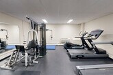 This fitness room at 935 Dundas St E in Mississauga, Ontario offers a practical and functional exercise space designed to accommodate a variety of workout routines. The room features a drop-panel ceiling with recessed fluorescent lighting that ensures consistent, even illumination throughout the space. Dark rubber flooring provides a durable, non-slip surface suited for active use.

The cardio section is well-appointed, featuring multiple treadmills, an elliptical trainer, and a stationary bike, giving users a range of aerobic options. A wall-mounted flat-screen television is positioned within view of the cardio equipment for entertainment during workouts. On the strength training side, a multi-station cable machine offers versatility for full-body resistance exercises, complemented by a weight bench for additional free-weight workouts.

A blue folding exercise mat is available for stretching and floor-based exercises, and a large wall mirror expands the sense of space while allowing users to monitor their form. The room includes a wall-unit HVAC system and baseboard heating to ensure year-round comfort. Practical amenities such as a paper towel dispenser, waste bin, and posted fitness guidelines reflect a well-maintained, user-focused environment. The space is accessed through a glass-paned door, adding a clean, professional finish to the overall layout.