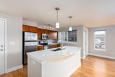 This well-appointed single-floor residence offers a thoughtfully designed open-concept layout that seamlessly connects the kitchen, dining, and living areas. The kitchen is a standout feature, showcasing rich cherry wood cabinetry, a subway tile backsplash, and a full suite of stainless steel appliances including a range, microwave, and refrigerator. A generously sized central island with a crisp white quartz countertop and an undermount sink serves as both a functional workspace and a natural gathering point, illuminated overhead by stylish pendant lighting. Warm hardwood flooring flows throughout the main living areas, complementing the neutral beige tones of the walls and creating a cohesive, inviting ambiance. Large windows on multiple sides of the unit flood the space with natural light while offering views of the surrounding urban landscape. The bathroom is equally refined, featuring a dual-sink vanity with white quartz countertops, rich wood cabinetry with brushed nickel hardware, a large wall mirror, and contemporary lighting. A carpeted bedroom benefits from a large window overlooking a landscaped courtyard, providing a quiet retreat. The overall design balances modern finishes with warm, natural materials, resulting in a comfortable and functional living space.