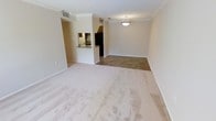 This single-floor residence offers a clean, open layout that seamlessly connects the living, dining, and kitchen areas. The spacious living area is finished with plush wall-to-wall carpeting, white walls, and elegant crown molding that runs throughout, lending the space a polished and cohesive aesthetic. A pendant light fixture hangs above the dining area, providing focused illumination and a subtle decorative touch.

The transition from the carpeted living space to the tiled dining and kitchen area is clearly defined, adding visual interest to the open floor plan. The kitchen is partially separated by a half-wall breakfast bar with a dark countertop, offering a convenient pass-through space ideal for casual dining or entertaining. Dark wood cabinetry provides ample storage, and a black refrigerator anchors the kitchen's functional design. A recessed sink area is visible just beyond the bar, completing the kitchen setup.

Ceiling vents are positioned throughout for efficient climate control, and the neutral color palette of warm beige tones across the walls, carpet, and tile creates a calm and inviting atmosphere. The overall ambiance is bright, clean, and move-in ready, making it an adaptable space suitable for a variety of living arrangements.