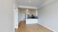 This single-floor space features an open-concept design that seamlessly connects the living area to the kitchen, creating a bright and functional environment. Light hardwood-style flooring extends throughout, contributing to a warm and cohesive aesthetic.

The kitchen is a standout feature, equipped with stainless steel appliances, including a refrigerator, microwave, and range. White cabinetry provides ample storage, while dark countertops offer a subtle contrast. A peninsula with a raised breakfast bar separates the kitchen from the living area, adding both utility and visual definition to the space.

The walls are finished in a neutral, light gray tone, complementing the overall modern aesthetic. A textured white ceiling adds subtle dimension overhead. The open corridor visible beyond the kitchen leads to additional rooms, each accessible via white panel doors, suggesting a practical and well-organized layout.

Natural and artificial lighting work together to keep the space feeling open and airy. The clean lines, neutral palette, and contemporary finishes throughout make this a versatile and welcoming space suited to a variety of uses.
