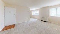 This 1-bedroom, 1-bathroom residence is designed with a straightforward, single-floor layout that maximizes functionality and ease of living. The living area is generously sized, featuring neutral-toned wall-to-wall carpeting and warm beige walls that create a clean, open atmosphere. Multiple windows fitted with horizontal blinds allow natural light to filter in, brightening the space throughout the day. An arched doorway adds a subtle architectural detail, connecting the living area to the rest of the unit. The kitchen is a standout feature, outfitted with rich dark wood cabinetry, dark granite-style countertops, and warm hardwood-style flooring. Black appliances — including a refrigerator, gas range, dishwasher, and microwave — provide a cohesive, functional aesthetic. Ample cabinet storage and counter space make this kitchen well-suited for everyday cooking. The bathroom is finished with floor-to-ceiling neutral tile surrounding a full soaking tub and shower combination, complemented by clean white fixtures and classic tile flooring. The bedroom offers a comfortable, carpeted retreat consistent with the overall neutral palette of the home. Together, the rooms form a cohesive, livable space with a clean and understated design throughout.