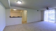 This single-floor unit offers a clean, functional layout designed for comfortable everyday living. The main living area is spacious and open, finished with plush wall-to-wall carpeting that adds warmth underfoot. A ceiling fan is mounted above, providing both comfort and light to the room. The smooth white walls and textured ceiling contribute to a bright, neutral atmosphere throughout.

The kitchen is positioned adjacent to the living area and is accessible through an open pass-through cutout, creating a connected feel between the two spaces. The kitchen features white upper cabinetry, dark countertops, and tile flooring, offering a practical contrast to the softer tones of the living area. A white refrigerator and standard appliances are visible, making it a fully equipped cooking space. Under-cabinet lighting illuminates the workspace effectively.

A doorway visible from the living area leads further into the unit, suggesting access to additional rooms beyond. The overall layout is straightforward and well-organized, with consistent neutral tones, recessed ceiling fixtures, and ample wall space throughout. This unit presents a tidy, move-in ready environment with a cohesive design that balances simplicity with functionality.
