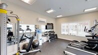 This fitness center offers a comprehensive workout environment designed to accommodate a variety of exercise routines. The space features durable speckled rubber flooring throughout, providing both safety and comfort underfoot. Neutral-toned walls and flush-mounted ceiling light panels contribute to a clean, functional aesthetic, while a wall-mounted split-system air conditioning unit ensures year-round climate control.

Cardio equipment is well-represented, with multiple Spirit-brand treadmills, an elliptical trainer equipped with its own monitor, and a recumbent stationary bike. A multi-function cable machine supports strength and resistance training, while a full dumbbell rack stocked with a wide range of weights caters to free weight workouts. Stability balls are also available for core and flexibility exercises.

A large wall-mounted flat-screen television serves as a central focal point, accompanied by a water cooler station for convenient hydration. Large windows fitted with white horizontal blinds line multiple walls, inviting ample natural light into the space and offering views of the surrounding exterior. A mirrored wall section enhances the sense of openness. Access to the room is provided through a windowed entry door, adding a layer of visibility and security to the space.