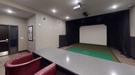 Located in Coon Rapids, MN, this single-level indoor golf simulator space is thoughtfully designed to deliver an immersive experience for golfers of all skill levels. The simulator bay itself is framed by floor-to-ceiling black drape curtains that enclose the hitting area, directing focus toward a large, bright projection screen mounted at the far end. A green artificial turf mat extends across the hitting zone, providing a realistic feel underfoot, while overhead sensors and a ceiling-mounted projector work together to capture and render each shot in vivid detail.

The viewing and lounge area behind the hitting mat is finished with a refined, club-like aesthetic. A long bar-height counter with a stone-look surface spans the back wall, paired with plush red leather swivel chairs that offer comfortable seating for players awaiting their turn. The dark wainscoting and muted green-gray wall tones of the lounge area create a sophisticated backdrop, accented by warm wall sconces and a decorative pendant light. A small media console with a monitor and beverage cooler sits conveniently at one end of the counter. Recessed lighting throughout ensures the entire space is well illuminated, contributing to a polished, welcoming atmosphere ideal for casual rounds or competitive play.