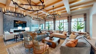 Located at 1905 Champs Dr in Naples, Florida, this sophisticated clubhouse offers a generously sized, single-floor layout designed for community gathering and social engagement. The grand entrance welcomes visitors with wide-plank hardwood flooring, a striking stone-patterned area rug, and dramatic chandelier lighting beneath coffered ceilings with warm wood beam accents. Multiple well-appointed hallways connect the various areas of the facility, featuring teal accent walls, curated artwork, glass-paneled doors, and elegant pendant lighting throughout. The expansive main living area is anchored by exposed wood beam ceilings, large-format chandeliers, bold navy built-in seating nooks with patterned wallpaper, and an abundance of natural light streaming through floor-to-ceiling black-framed windows. Dining areas include large communal tables set beneath woven rattan pendant lights atop polished stone-look tile flooring, complemented by multiple private rooms accessible through glass-paned doors. A distinguished reception area features soaring ceilings with wood beam detailing, a crystal chandelier, marble-look tile flooring, bold botanical wallpaper accents, and glass partition walls. The overall ambiance is coastal-contemporary, blending natural textures, curated furnishings, and vibrant design details throughout.