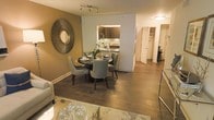 This single-floor residence showcases a thoughtfully designed open-concept layout that connects the living, dining, and kitchen areas into one cohesive and functional space. The dining area is a focal point, centered around a round dark-wood table paired with upholstered grey chairs, complemented by a striking sunburst mirror mounted on a warm taupe accent wall. Rich hardwood flooring flows throughout, adding warmth and continuity across the space.

The kitchen is partially open to the dining area, featuring dark cabinetry, a decorative tile backsplash, and stone countertops that contribute a refined aesthetic. The living area, partially visible at the forefront, includes a glass-topped coffee table and a plush area rug, reinforcing the space's contemporary character.

A hallway visible in the background leads to additional rooms, with doors suggesting well-organized private spaces, including what appears to be a closet with built-in storage. Recessed and track lighting provide layered illumination throughout. Overall, the space balances modern design with warm tones, creating an inviting and polished atmosphere suited for both everyday living and entertaining.