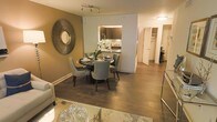This single-floor residence showcases a thoughtfully designed open-concept layout that connects the living, dining, and kitchen areas into one cohesive and functional space. The dining area is a focal point, centered around a round dark-wood table paired with upholstered grey chairs, complemented by a striking sunburst mirror mounted on a warm taupe accent wall. Rich hardwood flooring flows throughout, adding warmth and continuity across the space.

The kitchen is partially open to the dining area, featuring dark cabinetry, a decorative tile backsplash, and stone countertops that contribute a refined aesthetic. The living area, partially visible at the forefront, includes a glass-topped coffee table and a plush area rug, reinforcing the space's contemporary character.

A hallway visible in the background leads to additional rooms, with doors suggesting well-organized private spaces, including what appears to be a closet with built-in storage. Recessed and track lighting provide layered illumination throughout. Overall, the space balances modern design with warm tones, creating an inviting and polished atmosphere suited for both everyday living and entertaining.