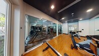 This expansive fitness center is thoughtfully designed to accommodate a wide range of workout preferences across two distinct zones. The main cardio and strength training area features patterned carpet flooring, exposed white ductwork, ceiling fans, and large floor-to-ceiling windows that flood the space with natural light and offer views of surrounding palm trees and greenery. This area is lined with mirrors along the walls, enhancing the sense of space, and is outfitted with treadmills, ellipticals, stair climbers, cable machines, strength training equipment, and a full dumbbell rack. A separate cycling studio, enclosed by full-height glass walls, features polished hardwood floors, a dramatic dark ceiling with recessed lighting, wall-mounted mirrors, a flat-screen TV, and a lineup of stationary bikes. The entry corridor is finished with warm wood-look tile flooring, built-in cabinetry with a cushioned bench, and a mounted display screen, creating a welcoming transition into the facility. Decorative angular ceiling fixtures add a contemporary design touch throughout. Large glass doors in the cardio zone open directly to an outdoor area, seamlessly connecting the indoor fitness experience with the exterior environment.
