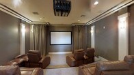 This dedicated home theater is thoughtfully designed to deliver an immersive cinematic experience. At the heart of the room is a large, centrally positioned projection screen framed by floor-to-ceiling drapes in a warm taupe tone, which line the walls and contribute to both the acoustic quality and the sophisticated ambiance of the space. A ceiling-mounted projector is positioned directly overhead, providing a clean and professional installation that keeps the focus on the screen. Recessed lighting is subtly integrated into the ceiling, complemented by wall sconces that offer soft, ambient illumination ideal for a theater setting. The seating arrangement consists of generously sized brown leather recliner chairs, arranged to ensure clear sightlines to the screen from every position. The neutral-toned carpeted flooring and muted wall colors work together to minimize light reflection and enhance the viewing environment. Crown molding adds a refined architectural detail to the ceiling perimeter, elevating the overall design aesthetic. The result is a well-appointed, purpose-built theater room that balances functionality with an upscale, comfortable atmosphere suited for both casual movie nights and more formal screenings.