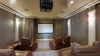 This dedicated home theater is thoughtfully designed to deliver an immersive cinematic experience. At the heart of the room is a large, centrally positioned projection screen framed by floor-to-ceiling drapes in a warm taupe tone, which line the walls and contribute to both the acoustic quality and the sophisticated ambiance of the space. A ceiling-mounted projector is positioned directly overhead, providing a clean and professional installation that keeps the focus on the screen. Recessed lighting is subtly integrated into the ceiling, complemented by wall sconces that offer soft, ambient illumination ideal for a theater setting. The seating arrangement consists of generously sized brown leather recliner chairs, arranged to ensure clear sightlines to the screen from every position. The neutral-toned carpeted flooring and muted wall colors work together to minimize light reflection and enhance the viewing environment. Crown molding adds a refined architectural detail to the ceiling perimeter, elevating the overall design aesthetic. The result is a well-appointed, purpose-built theater room that balances functionality with an upscale, comfortable atmosphere suited for both casual movie nights and more formal screenings.