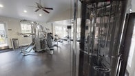 This fitness center is a thoughtfully designed, single-floor facility built to support a full range of workout routines. The space is divided into distinct functional zones, each oriented toward a specific type of training. The cardio area is positioned along a wall of large French doors and windows, flooding the zone with natural light and offering views of the surrounding outdoor area. It houses multiple treadmills and elliptical machines, complemented by wall-mounted televisions for entertainment during workouts. The strength training area features a multi-station cable and weight machine system, including Matrix-branded equipment, offering a wide variety of resistance exercises. A dedicated free weight section provides a tiered dumbbell rack stocked with a full range of weights, along with a flat bench for targeted exercises. Throughout the facility, dark rubber flooring provides a durable and safe surface, while ceiling fans help maintain comfortable airflow. The interior design incorporates a crisp white and navy blue color scheme, with recessed lighting and textured accent walls adding visual depth. Restroom facilities are conveniently located on-site. The space is bright, clean, and organized, creating a practical and motivating environment for fitness.