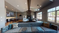 This elegantly designed single-floor common area space showcases a striking open-concept layout anchored by soaring vaulted ceilings that amplify the sense of grandeur throughout. The main lounge is a centerpiece of refined design, featuring a floor-to-ceiling dark-paneled fireplace surround paired with a bold circular ring chandelier that commands attention from above. Generous seating arrangements, including curved sectional sofas and a tufted accent sofa, are thoughtfully positioned to encourage both intimate conversation and relaxed gathering.

Warm hardwood flooring extends throughout, complementing the cool-toned grey walls and contemporary furnishings. A bank of large multi-pane windows floods the space with natural light while offering views of the outdoor grounds, including what appears to be a pool area and landscaped surroundings. French doors provide seamless access to additional spaces or outdoor areas.

A sleek wet bar and kitchenette area runs along one side of the room, complete with bar-height seating, pendant lighting, stone countertops, dark cabinetry, and curated wall art — making it well-suited for social gatherings. The overall ambiance strikes a balance between modern sophistication and welcoming comfort, with carefully chosen accent pieces in teal and navy adding cohesive pops of color throughout.