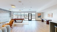 Located along the South Side Waterfront in Pittsburgh, this thoughtfully designed amenity space spans a generous single floor, offering a seamless blend of social, recreational, and functional areas. Light hardwood flooring flows throughout, tying each zone together with a clean, contemporary aesthetic.

The open living area serves as a central gathering hub, featuring a dramatic textured stone accent wall with a wall-mounted TV, stylish lounge seating, and recessed ceiling lighting. Adjacent to it, a dedicated game room is outfitted with a full-size billiard table and a shuffleboard table, bathed in natural light from expansive floor-to-ceiling windows that open directly to the outdoor terrace.

The dining area is equally refined, centered around a large oval dining table beneath a sculptural brass chandelier, with framed artwork adorning the walls and oversized glass doors providing direct terrace access. The kitchen impresses with striking terrazzo countertops and backsplash, professional-grade stainless steel appliances, white cabinetry, and a bold gold pendant light.

The wraparound outdoor terrace offers sweeping views of the Pittsburgh skyline and surrounding greenery, enclosed by sleek black metal railings. A separate media nook with a large-screen TV completes the space, making it a versatile and well-appointed environment for entertaining and community gatherings.