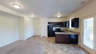 This single-floor space offers a practical and well-organized layout centered around an open-concept design that connects the kitchen and living area. The kitchen is a standout feature, equipped with rich dark espresso cabinetry that extends to the upper and lower levels, providing generous storage. Black appliances — including a refrigerator, electric range, built-in microwave, and sink — are neatly integrated into the layout. A peninsula countertop with a light stone-style surface extends outward, offering additional prep space and a natural division between the kitchen and the adjoining living area. The flooring transitions from tile in the kitchen zone to plush carpet in the living space, clearly delineating functional areas. Recessed flush-mount ceiling fixtures provide consistent overhead lighting throughout. White interior doors with raised panel detailing lead to additional rooms, maintaining a clean, cohesive aesthetic. The neutral wall tones and white ceilings contribute to a bright, airy atmosphere. A window near the sink allows natural light to filter into the kitchen, enhancing the overall sense of openness in this efficiently designed space.