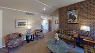 This single-floor clubhouse is thoughtfully designed to accommodate gatherings of all sizes, combining comfort and functionality throughout its open layout. The main lounge area serves as the heart of the space, anchored by a striking full-height brick fireplace that adds warmth and character. Vaulted ceilings with recessed lighting and a ceiling fan enhance the sense of openness, while plush seating arrangements — including sofas and upholstered chairs in classic patterns — create a relaxed, inviting atmosphere. Natural light filters in through multiple windows fitted with wooden blinds, brightening the carpeted interior.

Adjacent to the lounge, a formal dining area features vaulted ceilings accented by decorative crown molding and windows on multiple walls. A large wooden dining table with seating for several guests makes it well-suited for group meals or meetings, and a bold woven tapestry adds an artistic focal point.

The fully equipped galley-style kitchen is designed for practical use, offering generous counter space with teal laminate surfaces, white cabinetry, a full-size range, refrigerator, dishwasher, and multiple microwaves. The slate-tile flooring and patterned wallcovering give the kitchen a distinct, cozy character. A pass-through connects the kitchen directly to the dining area, ensuring a smooth flow for events and gatherings.