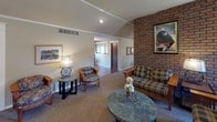 This single-floor clubhouse is thoughtfully designed to accommodate gatherings of all sizes, combining comfort and functionality throughout its open layout. The main lounge area serves as the heart of the space, anchored by a striking full-height brick fireplace that adds warmth and character. Vaulted ceilings with recessed lighting and a ceiling fan enhance the sense of openness, while plush seating arrangements — including sofas and upholstered chairs in classic patterns — create a relaxed, inviting atmosphere. Natural light filters in through multiple windows fitted with wooden blinds, brightening the carpeted interior.

Adjacent to the lounge, a formal dining area features vaulted ceilings accented by decorative crown molding and windows on multiple walls. A large wooden dining table with seating for several guests makes it well-suited for group meals or meetings, and a bold woven tapestry adds an artistic focal point.

The fully equipped galley-style kitchen is designed for practical use, offering generous counter space with teal laminate surfaces, white cabinetry, a full-size range, refrigerator, dishwasher, and multiple microwaves. The slate-tile flooring and patterned wallcovering give the kitchen a distinct, cozy character. A pass-through connects the kitchen directly to the dining area, ensuring a smooth flow for events and gatherings.
