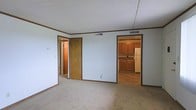 This single-floor residential space offers a straightforward and functional layout designed for comfortable everyday living. The main living area is finished with wall-to-wall carpet in a neutral tone, providing a soft and practical surface underfoot. Light blue walls and a textured drop ceiling lend the room a clean, understated look, while crown molding along the upper perimeter adds a subtle decorative touch. HVAC vents are positioned in the ceiling, ensuring climate control throughout the space. Two doorways open from the main living area — one leads to additional rooms within the unit, and the other provides a direct view into the kitchen. The kitchen features warm-toned wood cabinetry offering ample storage, a functional countertop workspace, and a full-size refrigerator. The kitchen floor is finished with a hard surface material, creating a practical transition from the carpeted living space. The overall ambiance of the unit is modest and move-in ready, with a neutral color palette that provides a versatile backdrop for a variety of furnishing styles. The open sightlines between rooms contribute to a connected and cohesive feel throughout the space.