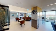Located at 125 E Washington St in the heart of downtown Phoenix, this upscale single-floor space offers a thoughtfully designed interior that blends contemporary architecture with high-end materials. The open-plan layout flows seamlessly between distinct functional zones, unified by polished light-toned flooring and crisp white ceilings with recessed and linear lighting throughout.

A central island bar serves as the focal point of the entertaining area, topped with dramatic veined stone and surrounded by richly upholstered rust-colored bar seating beneath an elegant sculptural pendant light fixture. Adjacent to the bar, a built-in wet bar features matching stone countertops, a wine cooler, under-counter refrigeration, and a sleek matte black sink — all framed by warm wood cabinetry and bold marble slab backsplash detailing.

A striking stone-clad column with gold-accented paneling anchors the main gathering area, which includes casual dining tables and plush lounge seating in jewel tones. Curated artwork adds personality throughout. Floor-to-ceiling glass walls and sliding doors open directly onto a spacious outdoor terrace, flooding the interior with natural light and offering sweeping views of the Phoenix skyline.