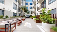 Located at 1430 Lincoln Blvd in Santa Monica, California, this contemporary multi-story residential building exemplifies modern coastal living. The building's clean, white stucco façade is accented by large windows and private balconies with sleek metal railings, offering residents both style and outdoor access.

At the heart of the property lies a spacious, open-air courtyard that serves as a central gathering space. The courtyard is thoughtfully designed with large-format light-colored pavers, creating a bright and welcoming atmosphere. Lush landscaping throughout includes tall palm trees, ornamental grasses, and oversized white and cream-toned planters, bringing natural vibrancy to the outdoor environment. Raised planter beds with warm Corten steel accents further enhance the landscaped areas.

Comfortable outdoor lounge seating with bold red-framed furniture provides inviting areas for residents to gather and socialize. The building's entry points are framed by decorative warm-toned slatted wood and metal fencing, adding a layer of privacy while maintaining an open, airy feel. Floor-to-ceiling glass at the main entrance allows natural light to flow through the lobby. The overall design seamlessly blends Southern California's indoor-outdoor lifestyle with sophisticated, contemporary architecture.