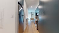 This thoughtfully designed single-floor space combines contemporary aesthetics with efficient use of layout throughout. The galley-style kitchen is a standout feature, outfitted with bold teal cabinetry, a mosaic tile backsplash, light quartz countertops, and a full suite of stainless steel appliances including a dishwasher, range, microwave, and a full-size refrigerator. Recessed lighting and light oak hardwood flooring run throughout, creating a cohesive, modern feel.

The living area is warmly appointed, featuring large windows dressed with roller shades and floor-length curtains that allow for flexible natural light control and views of the surrounding urban environment. A comfortable sofa, coffee table, and ambient floor and table lamps contribute to a relaxed, curated atmosphere. A framed art piece anchors the living wall, adding a refined touch.

The sleeping area is cleverly separated from the living space by a floor-to-ceiling curtain panel, providing privacy without enclosing the area entirely, maintaining an open and airy flow. The bathroom features clean white square tile, a glass-enclosed walk-in shower with chrome fixtures, and tile flooring, offering a crisp, functional finish throughout.