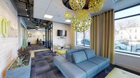 This single-floor space showcases a thoughtfully designed interior that blends contemporary style with functional versatility. The lounge area serves as a focal point, anchored by a plush blue sofa and complemented by accent chairs in a coordinating palette. Overhead, striking yellow-green spherical pendant lights with an open lattice design draw the eye upward and infuse the space with artistic energy.

Natural light filters in through large windows dressed with floor-length neutral curtains, offering views of the surrounding exterior. A wall-mounted flat-screen television is positioned above a light wood accent wall, creating a media-ready nook ideal for informal gatherings or presentations.

The flooring features patterned commercial carpet in a grey geometric design that grounds the space visually. Glass-partitioned walls separate the lounge from adjacent work areas, maintaining an open, airy feel while defining distinct zones for focused work. Beyond the partitions, individual workstations are visible, suggesting a professional coworking or office environment. The overall ambiance is energetic yet polished, balancing creative design with practical workplace functionality.