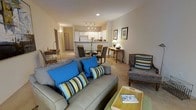 This single-floor space showcases an open-concept layout that seamlessly connects the living room, dining area, and kitchen into one cohesive, functional environment. The living room serves as the centerpiece, anchored by a plush gray sofa accented with vibrant blue and striped throw pillows, complemented by an upholstered accent chair. A light wood coffee tray table adds a casual, practical touch to the seating arrangement. Recessed and track lighting throughout provides consistent, warm illumination across the space. The dining area features a glass-top table surrounded by dark wood chairs, with a floral centerpiece adding a decorative accent. Adjacent to the dining space, a small console table with a lamp and decorative objects offers additional character. The kitchen is positioned at the rear of the open plan, featuring white cabinetry, white appliances, and a breakfast bar that visually separates it from the living and dining zones. The flooring transitions between tile and carpet, defining different functional areas within the open layout. Neutral beige walls and warm lighting contribute to a comfortable, welcoming ambiance throughout.
