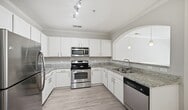 This 690-square-foot, one-bedroom, one-bathroom apartment — Design 1I #568 — is located at 141 Park at North Hills St in Raleigh, NC. The open-concept kitchen and living area form the heart of the home, featuring light hardwood-style flooring, crown molding, and a ceiling fan throughout. The kitchen is well-appointed with white shaker cabinetry, granite countertops, and a full suite of stainless steel appliances including a refrigerator, electric range, and built-in microwave. The living room is bright and airy, highlighted by French doors with transom windows that invite natural light and provide balcony access. The 180-square-foot primary bedroom is finished with the same hardwood-style flooring, crown molding, a ceiling fan, and two closet doors for ample storage. The bathroom features a full tub-shower combo with white subway tile surround, a white vanity with modern hardware, and coordinating flooring. A dedicated laundry area houses a stacked washer and dryer unit. Neutral warm-gray walls and clean white trim create a cohesive, modern aesthetic throughout the entire apartment.