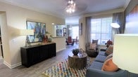 This thoughtfully designed single-floor residence offers a seamless blend of comfort and sophistication. The open-concept layout connects the living and dining areas, creating a spacious and inviting atmosphere ideal for both relaxing and entertaining. The living area is anchored by a plush sectional sofa, a rustic round wooden coffee table, and a vibrant geometric area rug that adds a pop of color to the warm, neutral palette. An eye-catching chandelier serves as a focal point overhead, complementing the crown molding that frames the room with a refined architectural touch. Hardwood-style flooring runs throughout, adding warmth and continuity across the space. A large window dressed with elegant drapes floods the area with natural light, offering a bright and airy feel. A sleek black credenza with decorative accents and bold artwork anchors one wall, adding a contemporary artistic element. A pass-through opening connects the living space to the dining area, where a pendant light and coordinating furniture continue the cohesive design aesthetic. The overall ambiance is modern yet welcoming, with carefully curated décor that balances style and livability throughout every corner of this well-finished home.
