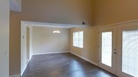 This two-story space makes a strong first impression with its dramatically vaulted ceilings that rise well above standard height, creating an expansive, open atmosphere. The main living and dining area showcases rich, dark hardwood flooring that runs throughout, providing a warm contrast against the neutral beige walls and bright white trim and baseboards. A classic chandelier with multiple light fixtures serves as a focal point overhead, adding a touch of elegance to the dining area. Natural light flows generously into the space through multiple windows fitted with white horizontal blinds, as well as a glass-paneled door that leads to the exterior, further enhancing the bright and welcoming ambiance. The vaulted ceiling design creates a split-level visual effect, with an elevated portion transitioning into a defined lower-ceiling zone, adding architectural interest and dimension. The ceiling-mounted HVAC vent indicates a centralized climate control system. Overall, the space maintains a clean, neutral palette that provides a versatile foundation, combining practical features with an open, light-filled character suited to a variety of uses.