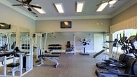 This fitness center offers a comprehensive workout environment designed to support a wide range of exercise routines. The space is thoughtfully laid out across a single floor, providing ample room to move freely between stations without feeling cramped. The walls are finished in a neutral light gray tone, complemented by white crown molding and baseboards that lend a clean, polished look to the interior. Recessed panel lighting and ceiling fans are evenly distributed overhead, ensuring the space stays well-lit and comfortably ventilated during use. Large mirrors line the walls, enhancing the sense of openness while allowing users to monitor their form. The equipment selection is diverse and includes a multi-function cable machine, a seated weight stack machine, a dumbbell rack with a full range of free weights, adjustable benches, and a dedicated press station. Additional cardio and functional training equipment is visible along the perimeter. A wall-mounted flat-screen television is positioned for easy viewing from multiple workout stations. The flooring consists of durable, low-pile carpeting suited for a gym environment. A private door provides a separate entry or exit point, contributing to the overall functionality of the space.