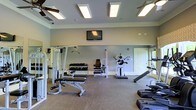 This fitness center offers a comprehensive workout environment designed to support a wide range of exercise routines. The space is thoughtfully laid out across a single floor, providing ample room to move freely between stations without feeling cramped. The walls are finished in a neutral light gray tone, complemented by white crown molding and baseboards that lend a clean, polished look to the interior. Recessed panel lighting and ceiling fans are evenly distributed overhead, ensuring the space stays well-lit and comfortably ventilated during use. Large mirrors line the walls, enhancing the sense of openness while allowing users to monitor their form. The equipment selection is diverse and includes a multi-function cable machine, a seated weight stack machine, a dumbbell rack with a full range of free weights, adjustable benches, and a dedicated press station. Additional cardio and functional training equipment is visible along the perimeter. A wall-mounted flat-screen television is positioned for easy viewing from multiple workout stations. The flooring consists of durable, low-pile carpeting suited for a gym environment. A private door provides a separate entry or exit point, contributing to the overall functionality of the space.