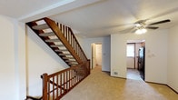 This three-floor home is thoughtfully laid out to maximize functionality across each level. The main living area features warm beige carpeting and neutral walls, creating a clean, understated backdrop. A prominent staircase with rich wood banisters and balusters connects the floors, serving as a central architectural element throughout the home. A ceiling fan overhead adds a practical touch to the main living space, which also benefits from natural light through multiple windows dressed with blinds.

The kitchen is equipped with wood cabinetry, dark stone-patterned countertops, and a door with a window that provides an exterior view, offering both storage and natural light. The bathrooms include a full bath with a tub-shower combo, tile flooring, and a wood-trimmed vanity, as well as a second bathroom featuring a sink, large mirror, and brushed nickel fixtures with textured walls.

The lower level includes an attached garage with a concrete floor and overhead door, as well as a dedicated laundry area featuring a washing machine, utility sink, and exposed plumbing along concrete block walls — a practical utility space that completes the home's multi-level layout.