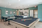 Located at 109 Cottage Grove Rd in Madison, Wisconsin, this well-appointed communal space offers a thoughtfully designed open-plan layout that seamlessly blends a fully equipped kitchen, a comfortable lounge area, and a recreational zone. The interior is distinguished by a consistent palette of soft teal-green walls, light hardwood flooring, and recessed lighting that together create a bright, welcoming ambiance throughout.

The kitchen is a standout feature, boasting a large central island with a white quartz countertop and seating for multiple guests on velvet navy barstools. White shaker cabinetry with brass hardware, stainless steel appliances, and a set of globe pendant lights with brass accents give the kitchen a clean, modern aesthetic that is both functional and visually appealing.

The living area is generously proportioned, furnished with a plush sectional sofa, leather accent chairs, and a glass coffee table arranged around a patterned area rug. A wall-mounted flat-screen television is positioned above a sleek white console table, making it an ideal spot for group gatherings. An elegant drum pendant light anchors the space with a refined, contemporary touch.

A pool table adds a recreational dimension, while a marble-top dining table with stylish chairs completes the social atmosphere. Large windows invite natural light and provide views of the surrounding greenery, enhancing the overall openness of this versatile space.