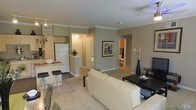 This single-floor residence offers an open-concept design that integrates the kitchen, dining, and living spaces into one cohesive area. The living room serves as the central gathering space, furnished with a cream-toned sofa set arranged around a dark coffee table atop plush carpeting. Crown molding lines the ceiling throughout, adding a refined architectural detail, while a ceiling fan provides functional comfort. The kitchen is efficiently laid out with light wood cabinetry, a full-size refrigerator, and a raised breakfast bar with bar stools, making it ideal for casual dining. Decorative elements, including a tall black vase with dried branches and framed wall art, contribute to a polished aesthetic. A hallway visible from the living area leads to additional rooms, suggesting a practical and well-organized floor plan. The flooring transitions from carpet in the living area to hard flooring in the kitchen and entryway zones. Neutral wall tones in warm gray create a cohesive backdrop throughout, allowing furnishings and accents to stand out. Overall, the space presents a comfortable, modern ambiance suited for everyday living.