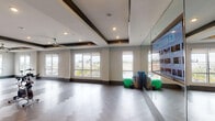 This well-appointed fitness studio offers a bright and open layout designed to support a variety of workout activities. The space features elegant herringbone-patterned hardwood flooring throughout, providing both aesthetic appeal and a durable surface suited for cycling, yoga, stretching, and group exercise classes. Multiple spin bikes are positioned across the open floor, offering ample room for group cycling sessions without feeling crowded. A full wall of floor-to-ceiling mirrors reflects the spacious interior and enhances the sense of openness, while a large mounted display screen provides access to on-demand workout content, making guided exercise sessions convenient and engaging. The ceiling design incorporates distinctive dark wood beam accents framing a tray ceiling with recessed lighting and multiple ceiling fans, ensuring the space remains well-lit and well-ventilated during intense workouts. Large windows line the walls, flooding the studio with natural light and offering views of the surrounding area. A TRX suspension training system and a rack of additional equipment are also available along one wall. Decorative abstract artwork adds a modern aesthetic touch to the environment. The overall ambiance is clean, contemporary, and energizing, making this studio a fully equipped and inviting space for fitness enthusiasts.