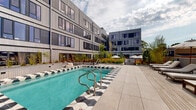 Located at 1111 Fairfield Ave in Cleveland's vibrant Ohio City neighborhood, this thoughtfully designed outdoor amenity space offers a resort-inspired experience within a contemporary residential setting. The centerpiece is a striking outdoor pool, framed by bold geometric black-and-white tiled coping that lends a distinctive, design-forward aesthetic. Adjacent to the pool, a generous sundeck is lined with lounge chairs and umbrellas, ideal for relaxing in open air.

The expansive wooden terrace extends across two connected zones, finished in light-toned hardwood decking that creates a warm, cohesive atmosphere. Curved white-paneled planters anchor the space with lush ornamental grasses, wispy trees, and seasonal plantings that add texture and color throughout. Multiple seating arrangements — including dining tables, lounge chairs, and casual bean bag seating — provide versatile settings for both social gatherings and quiet retreat.

Shaded umbrella clusters define intimate outdoor dining and lounging nooks, while built-in benches along planted walls offer additional casual seating. The surrounding multi-story building, clad in modern grey paneling with expansive windows, encloses the courtyard and provides a sense of privacy. Open skies above and abundant greenery throughout create a refreshing urban sanctuary well-suited for everyday outdoor living.