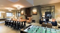 This single-floor amenity lounge is thoughtfully designed to blend comfort, style, and entertainment into one cohesive space. The room's centerpiece is a striking floor-to-ceiling stacked stone accent wall housing a linear fireplace, flanked by warm-toned artwork that adds a sophisticated, gallery-like feel. Warm ambient lighting, including a modern pendant fixture above the dining area, complements the earthy tones of the stone and wood furnishings throughout.

The dining area features a sturdy wooden table paired with contemporary stools, making it well-suited for casual gatherings or shared meals. Adjacent to the fireplace, a comfortable seating arrangement with upholstered chairs and a plush sofa invites residents to relax in a lounge-like atmosphere. Large windows along the perimeter allow natural light to filter in, enhancing the open and welcoming ambiance.

A foosball table anchors the recreational zone, adding a fun, social dimension to the space. A large framed mirror on one wall amplifies the sense of depth, while a coffee station or bar area is visible in the background, providing added convenience. The overall design strikes a balance between elegance and approachability, making this lounge an ideal gathering place for socializing and leisure.